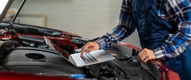 Kadın oto tamircisi dizüstü bilgisayar kullanarak motor kontrolü yapıyor.