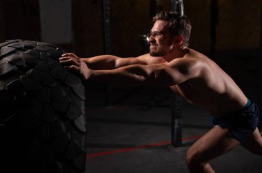Beyaz bir adam spor salonunda araba lastiği itiyor.