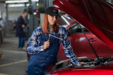 Kadın bir oto tamircisi arabanın açık kaputunda duruyor ve elinde bir tornavida tutuyor.