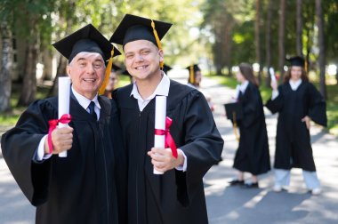 Bir grup mezun açık havada bornozla geziyor. Yaşlı bir adam ve genç bir adam diploma aldıkları için birbirlerini kutluyorlar.