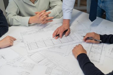 A caucasian man is standing making changes to a drawing, three colleagues are sitting at a table and listening to him