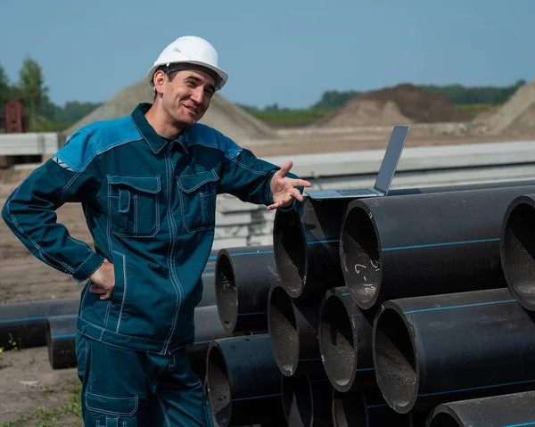 Stock image Caucasian male builder in a hard hat stands near the pipes and uses a laptop at a construction site