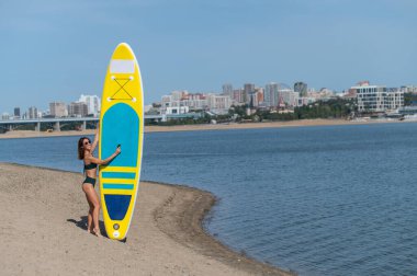 Beyaz bir kadın sahilde yürüyor ve şehirdeki nehirde bir yiyecek panosu taşıyor. Yaz sporu