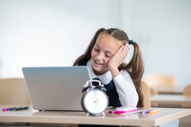 Beyaz kız okulda bir masada oturuyor ve bir dizüstü bilgisayarda okuyor. Masadaki çalar saat.