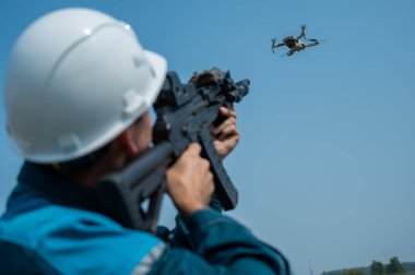 Kasklı beyaz bir adam tüfekle uçan bir dronu vuruyor.