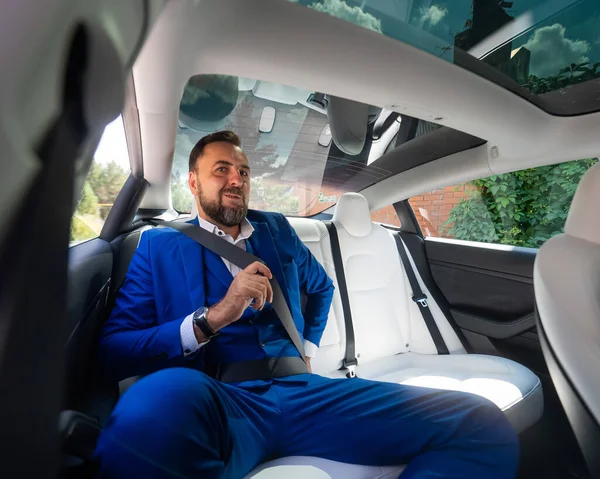 A caucasian man in a blue suit fastens his seat belt in the back seat of a car. Business class passenger