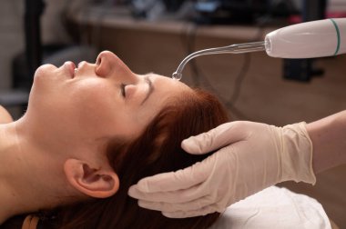 Doktor, kadın bir hastanın yüzündeki sivilce için darsonval aparatı kullanıyor.