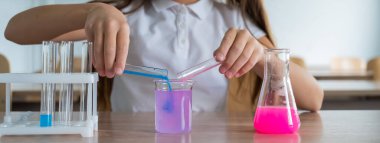 Liseli bir kız kimya dersinde deneyler yapıyor. Beher kabından renkli sıvılar döken bir kız.