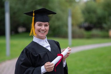 Mezuniyet elbisesi giymiş, açık havada diploma taşıyan mutlu beyaz kadın portresi.