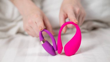 Yatakta uzanmış pembe ve mor vibratörler tutan bir kadın.