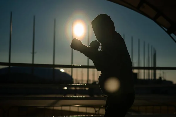 stock image A man in a sweatshirt trains in boxing at the stadium at sunset. Athlete silhouette