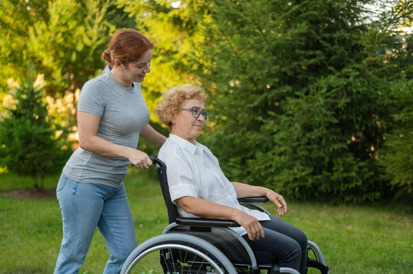 Biała Kobieta Niesie Starszą Matkę Siedzącą Wózku Inwalidzkim Przez Park — Zdjęcie stockowe