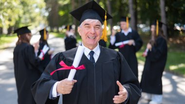 Bir grup mezun açık havada bornozla geziyor. Yaşlı bir öğrenci diploma aldığına sevinir.