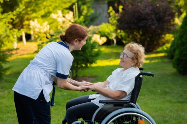 Bir hemşire, desteklemek için tekerlekli sandalyedeki yaşlı beyaz bir kadını elinden tutuyor. Hemşire bir hastayla parkta yürüyor.
