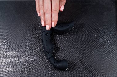 Bir kadın siyah bir prostat uyarıcısı tutar ve bu simülatör suyun yüzeyinde siyah bir zemin üzerinde dalgalanmalar yaratır.