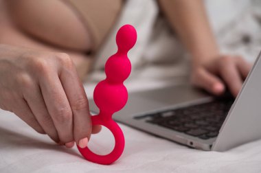 Yatakta yatarken dizüstü bilgisayarın yanında pembe anal boncuklar tutan kadın.