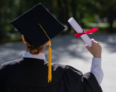 Rear view of a caucasian woman in a graduate gown holding a diploma outdoors