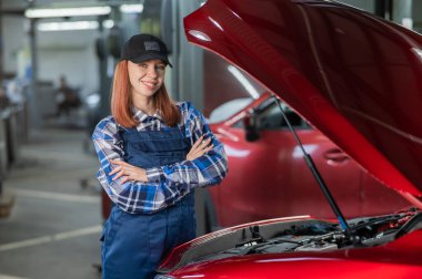 Otomobil tamircisi bir kadın, kollarını göğsüne bağlamış bir arabanın açık kaputunda duruyor.