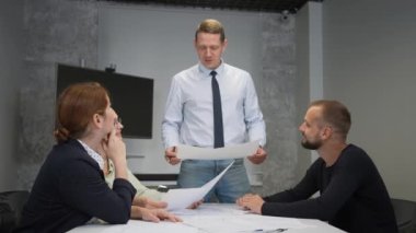 A caucasian man stands and holds a drawing, three colleagues sit at a table and listen to him. Slow motion