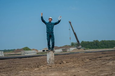 Koruyucu miğferli bir inşaatçı dizüstü bilgisayar tutuyor ve inşaat alanının ortasında bir yığın üzerinde duruyor.