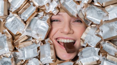 The face of a Caucasian woman surrounded by candy wrappers. The girl eats a bar of chocolate