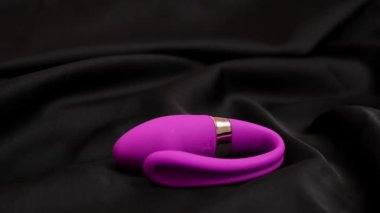 Bir kadın elini siyah ipek bir battaniyenin altından uzatır ve mor bir klitoral-vajinal seks oyuncağı alır.