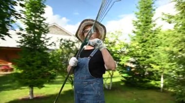 Şapkalı ve tulumlu yaşlı bir kadının portresi bir bahçe aleti tutuyor. Video 360 derece