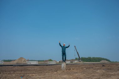 Koruyucu miğferli bir inşaatçı dizüstü bilgisayar tutuyor ve inşaat alanının ortasında bir yığın üzerinde duruyor.