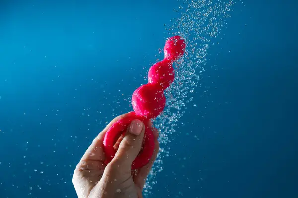 stock image Woman holding pink anal beads under running water on blue background. Sex toy hygiene concept