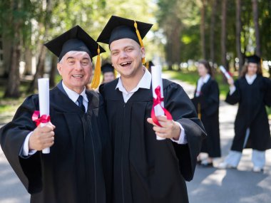 Bir grup mezun açık havada bornozla geziyor. Yaşlı bir adam ve genç bir adam diploma aldıkları için birbirlerini kutluyorlar.