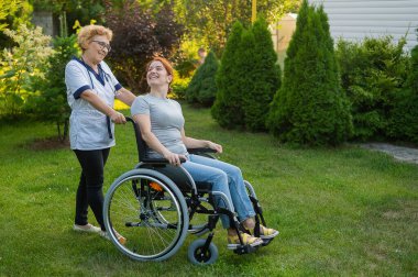 An elderly female nurse walks with a middle-aged woman in a wheelchair in the park