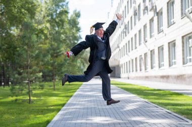 Mezuniyet elbisesi giymiş, dışarı atlayan ve diploması olan yaşlı mutlu bir adam.