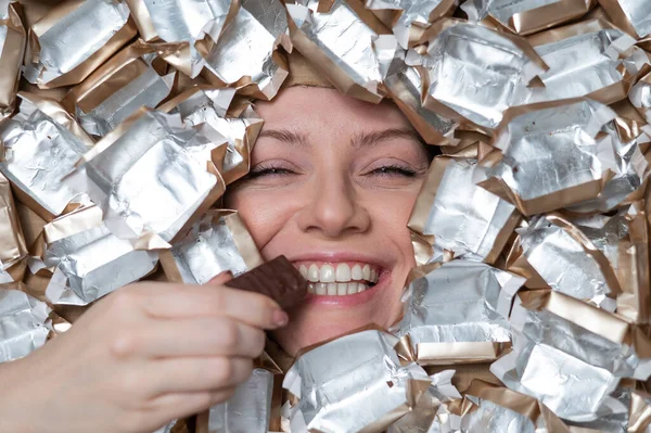 The face of a Caucasian woman surrounded by candy wrappers. The girl eats a bar of chocolate
