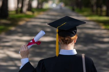 Mezuniyet elbisesi giymiş, elinde diplomasıyla üniversitenin yanında duran beyaz bir kadının arka görünüşü.