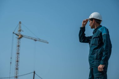 İş kıyafetleri ve kask giymiş bir müteahhit inşaat sahasının arka planında duruyor.