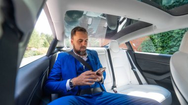 Mavi takım elbiseli beyaz bir adam arabanın arka koltuğunda akıllı telefon kullanır. Business class yolcusu