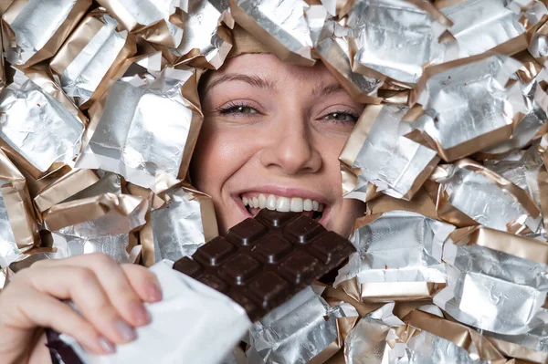 The face of a Caucasian woman surrounded by candy wrappers. The girl eats a bar of chocolate