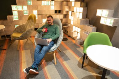 Caucasian bearded man sits in a comfortable chair drinks coffee and works on a laptop. Freelancer