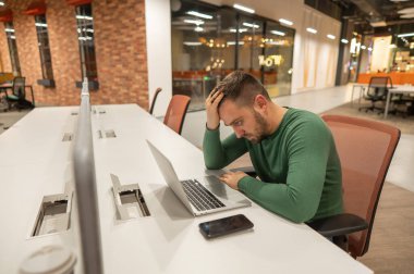 Açık bir uzay ofisinde dizüstü bilgisayarla çalışırken kafasını tutan sakallı bir adam.