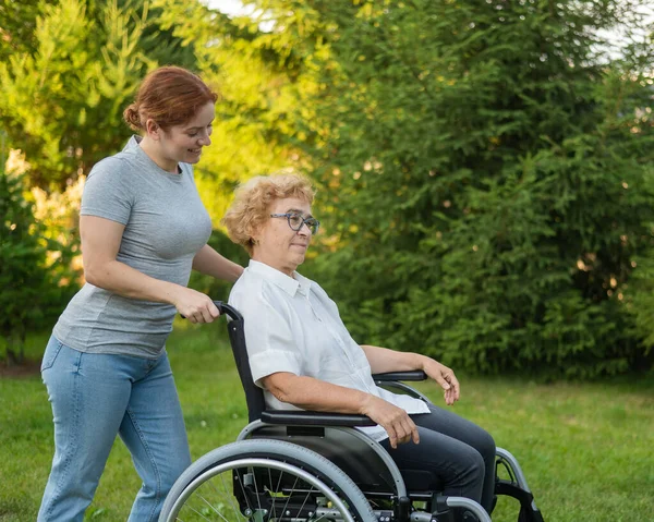 一位高加索妇女背着一位坐在轮椅上的老年母亲穿过公园 出去走走 — 图库照片