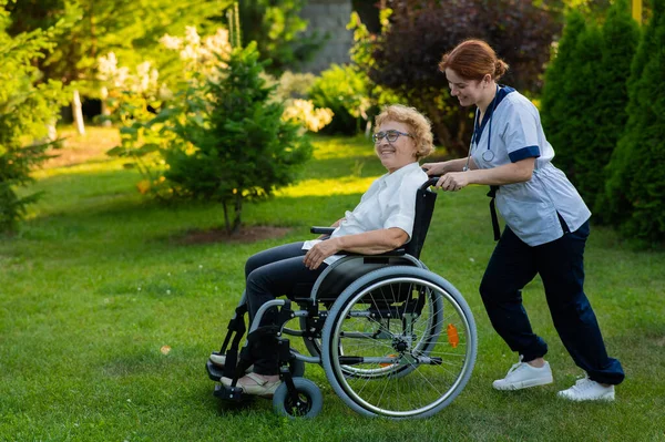 白人女医生和一位坐在轮椅上的老年病人在公园里散步 护士陪着一位老妇人在户外散步 — 图库照片