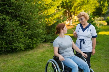 An elderly female nurse walks with a middle-aged woman in a wheelchair in the park