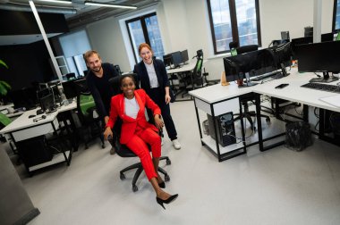 Kafkasyalı kızıl saçlı kadın, sakallı beyaz adam ofis koltuğunda Afro-Amerikan genç bayanı yuvarladı. Meslektaşlar iş yerinde eğleniyor.