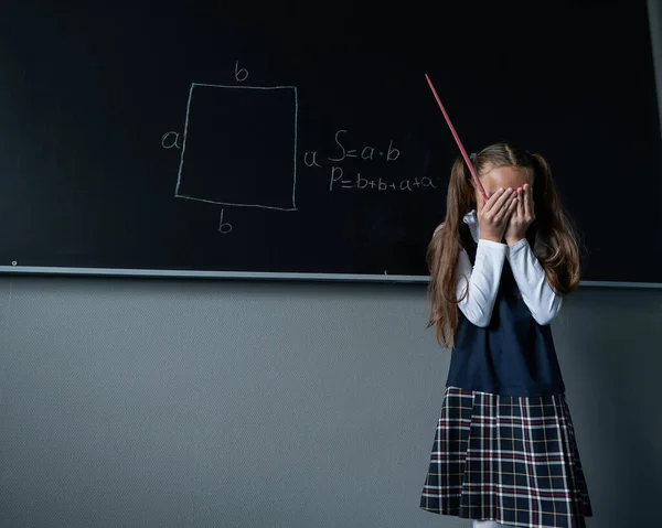 Caucasian girl stands at the blackboard and cries covering her face with her hands. The schoolgirl did not prepare for the lesson