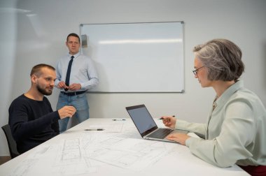 Beyaz tahtaya beyaz bir adam çiziyor, meslektaşları masaya oturuyor ve onu dinliyor.