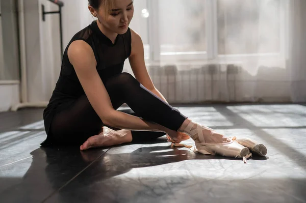 An Asian ballerina sits on the floor and puts on pointe shoes