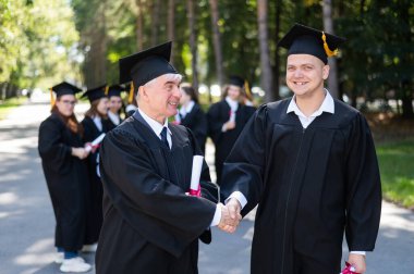 Bir grup mezun açık havada bornozla geziyor. Yaşlı bir adam ve genç bir adam diploma aldıkları için birbirlerini kutluyorlar.