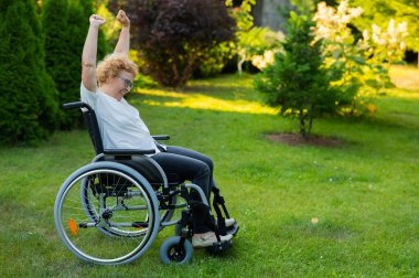 Elderly woman stretches her arms up while sitting in a wheelchair on a walk outdoors