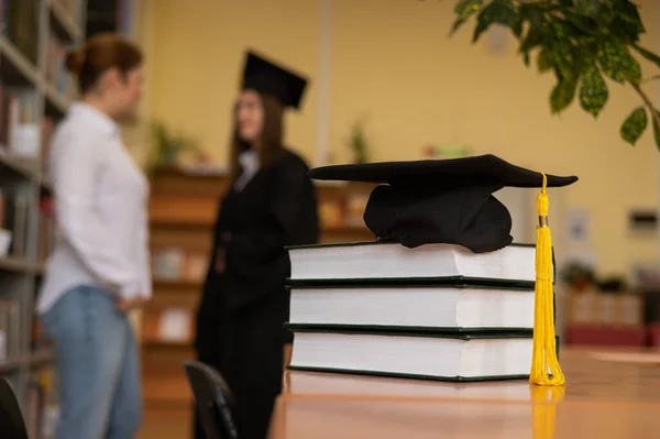 Close-up of a graduation cap on a stack of books in a library. teacher and young female graduate in the background