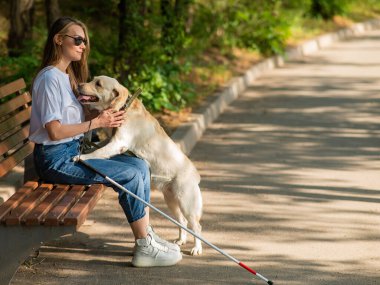 Kör genç bir kadın bankta otururken rehber köpeğe sarılıyor.